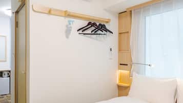 A hotel room with a bed, a wall-mounted air conditioner, a shelf with a spray bottle, and a window with curtains.