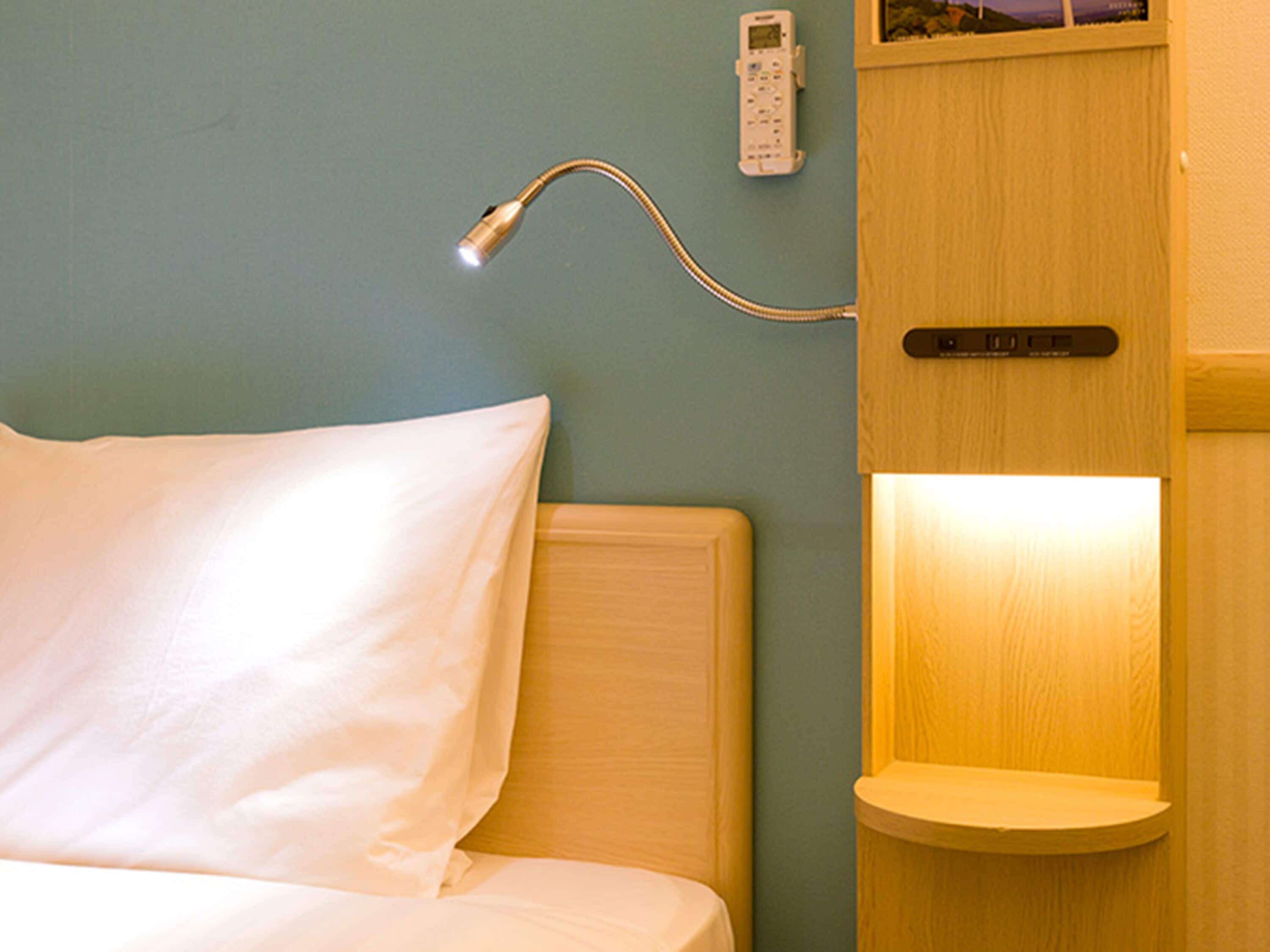 Hotellihuone, jossa on sänky, yöpöytälamppu, ohjauspaneeli ja pieni hylly.