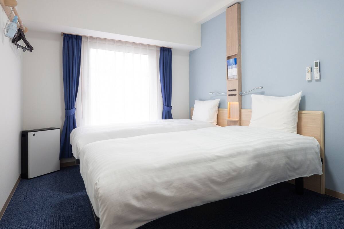 A hotel room with a large bed, a bedside table, a nightstand, a wall-mounted heater, and a window with blue curtains.