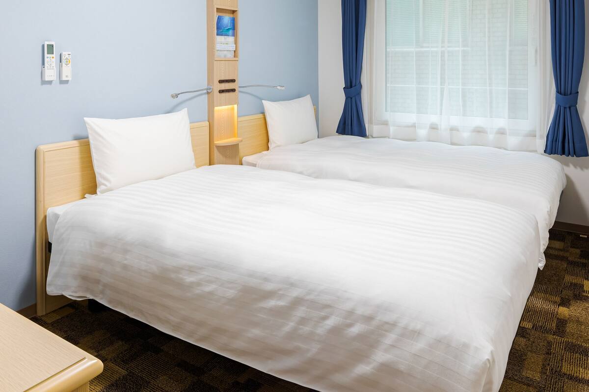 A hotel room with two beds, a wooden headboard, a bedside table, and a window with curtains.