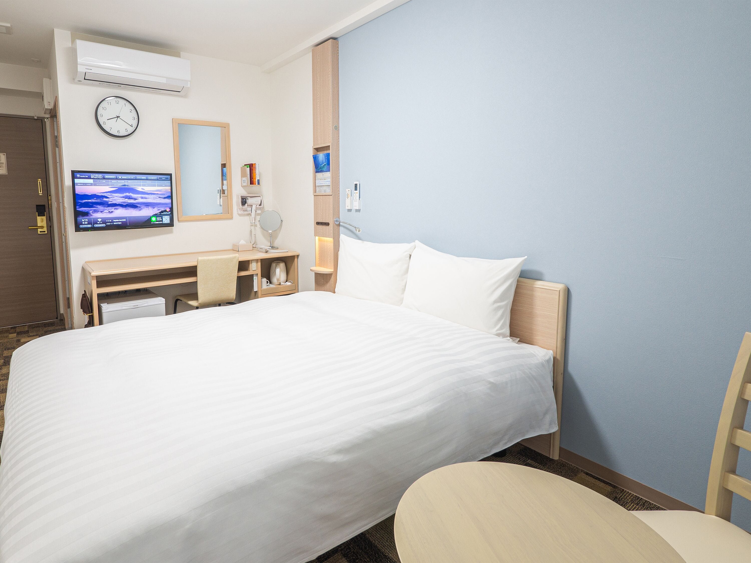 A hotel room with a large bed, a desk with a chair, a television, and a clock on the wall.