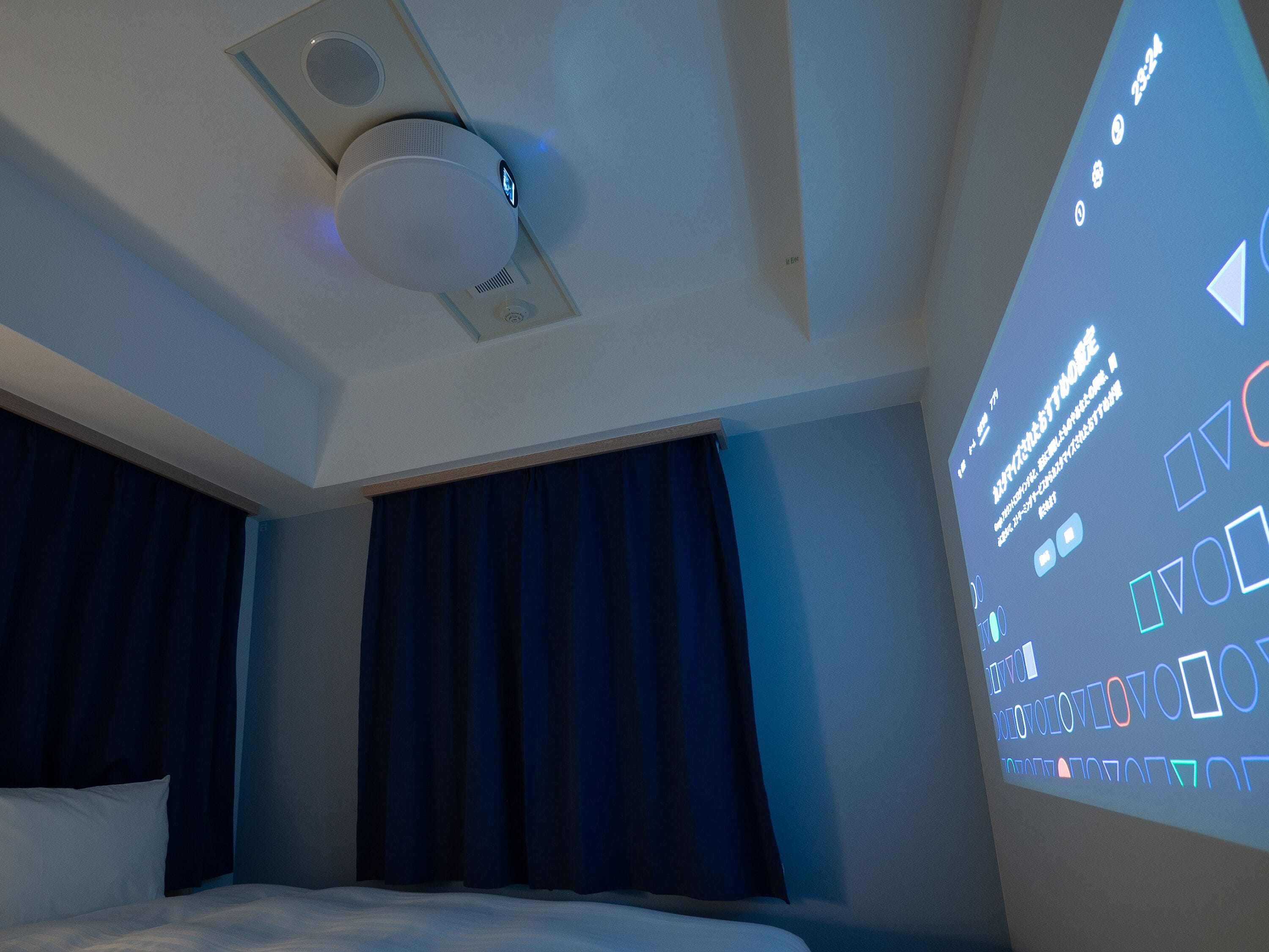 Una camera d'albergo con un letto, un rilevatore di fumo a soffitto e uno schermo di proiezione che mostra del testo.