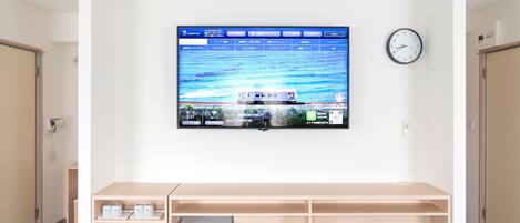 A hotel room with a large flat-screen TV mounted on the wall displaying a scenic image, a wooden desk with a small refrigerator, a blue stool, and a clock on the wall.