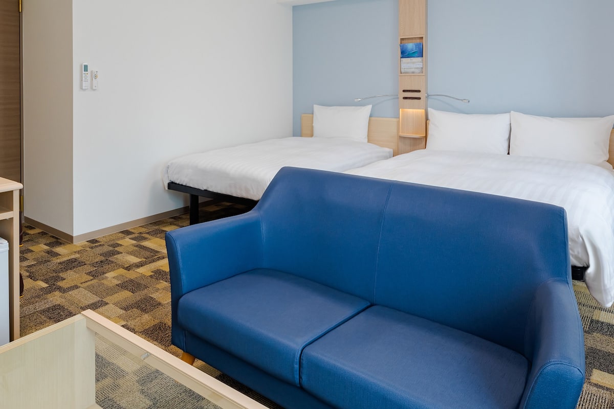 A hotel room with two beds, a blue sofa, a wooden wardrobe, and a wall-mounted control panel.