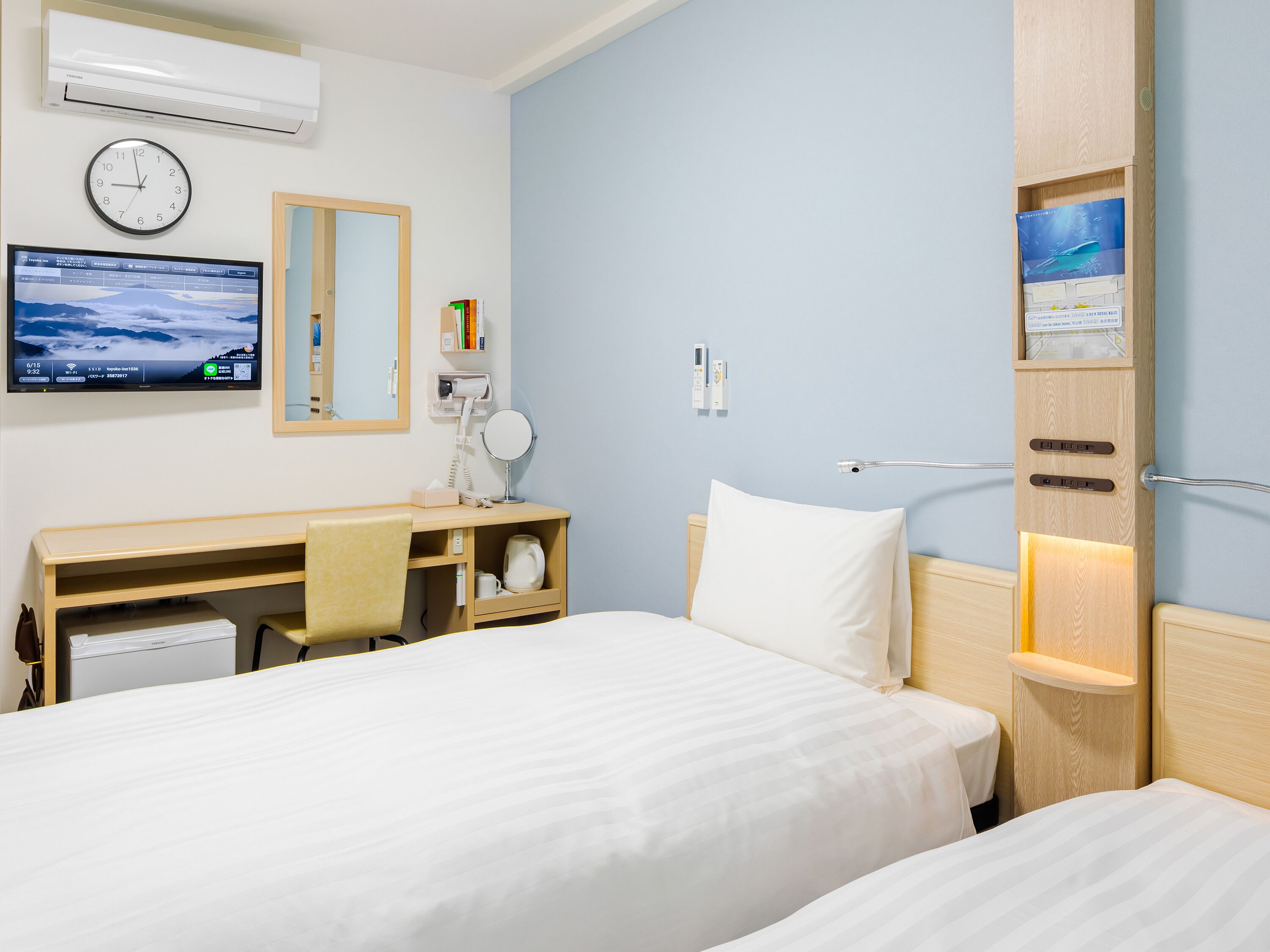 Habitación de hotel con dos camas, un escritorio con computadora, un reloj y un televisor de pared.
