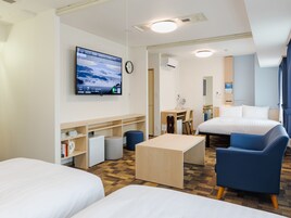 A hotel room with two beds, a desk, a chair, a TV, and a small kitchenette.