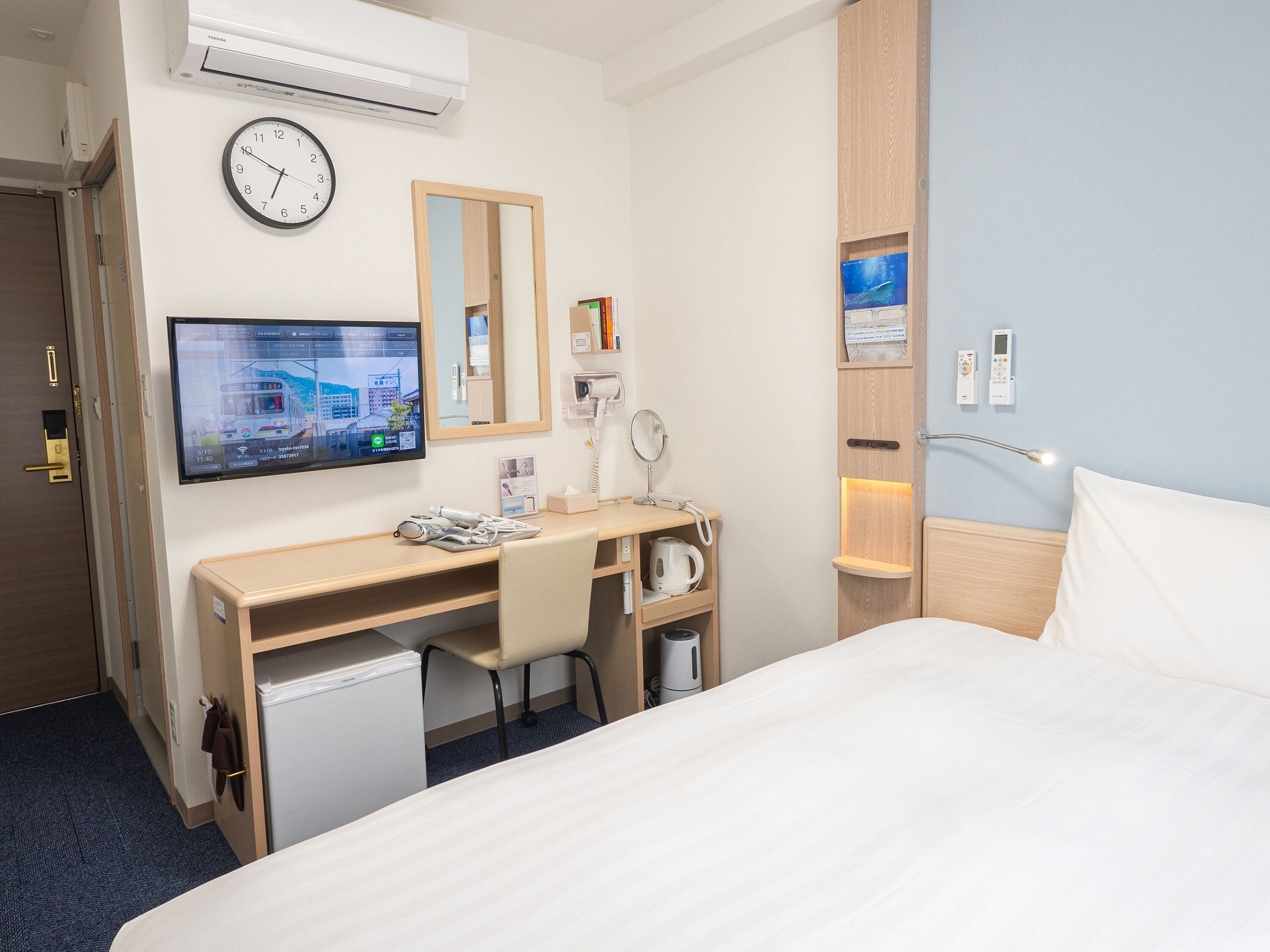 A hotel room with a bed, a desk with a computer, a clock, and a mirror.