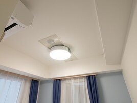 Quarto com ar-condicionado montado no teto, iluminação embutida e janela com cortinas translúcidas e azul-escuro.