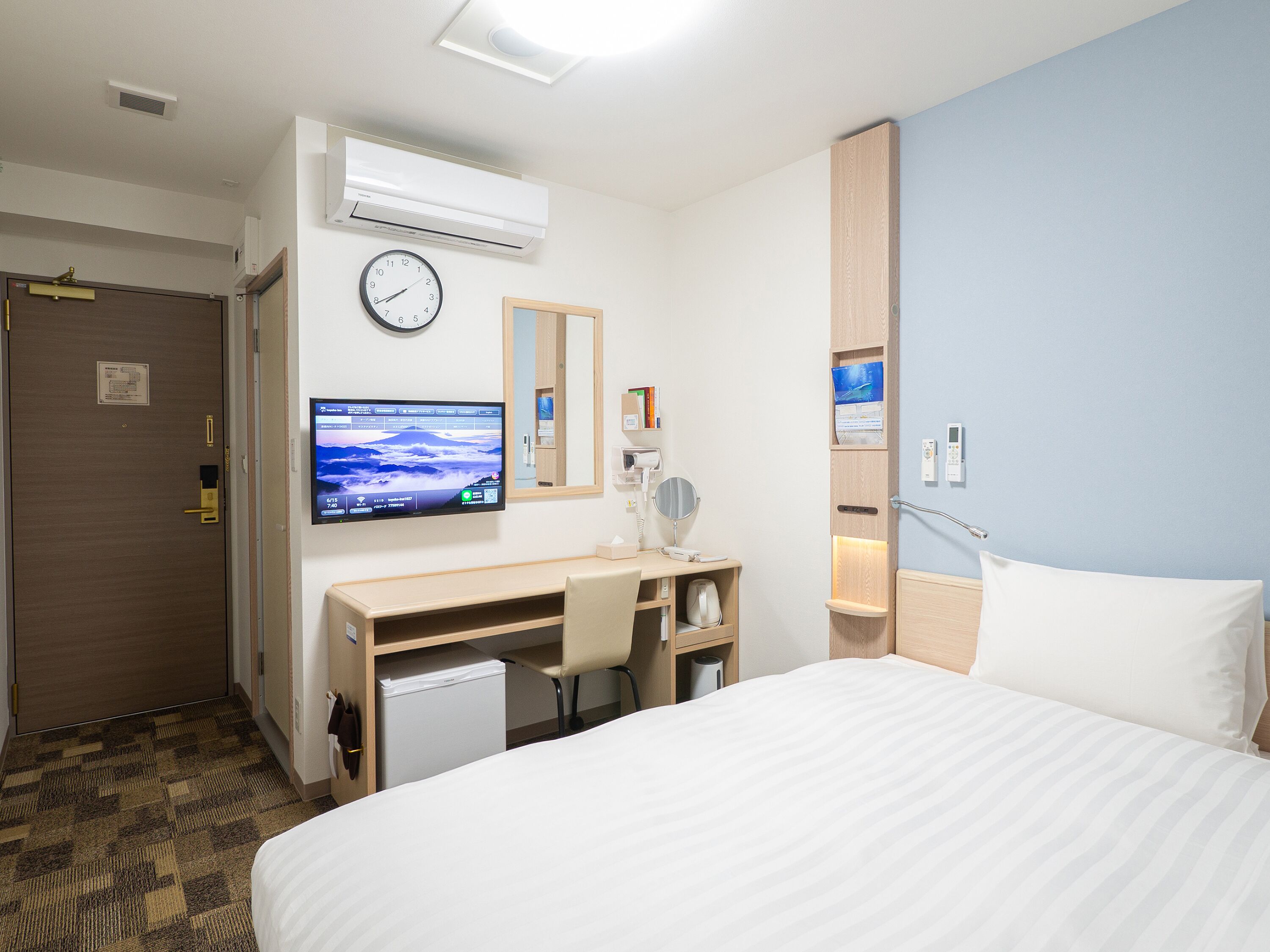 Habitación de hotel con cama, escritorio con silla, televisión, reloj y una pequeña estantería con libros y espejo.