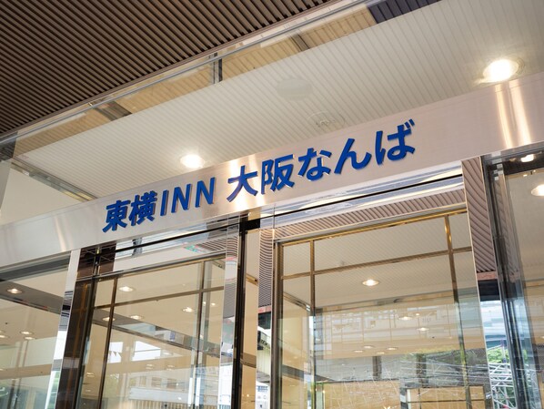 A hotel entrance with a sign in Japanese and English, large glass doors, and a ceiling with recessed lighting.