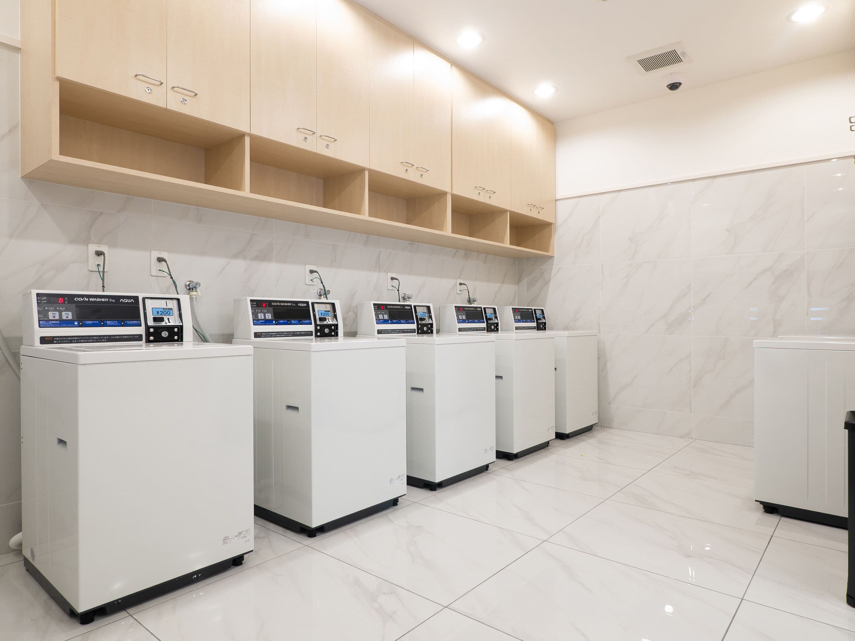A row of white laundry machines with control panels, wooden cabinets above, and a tiled floor.