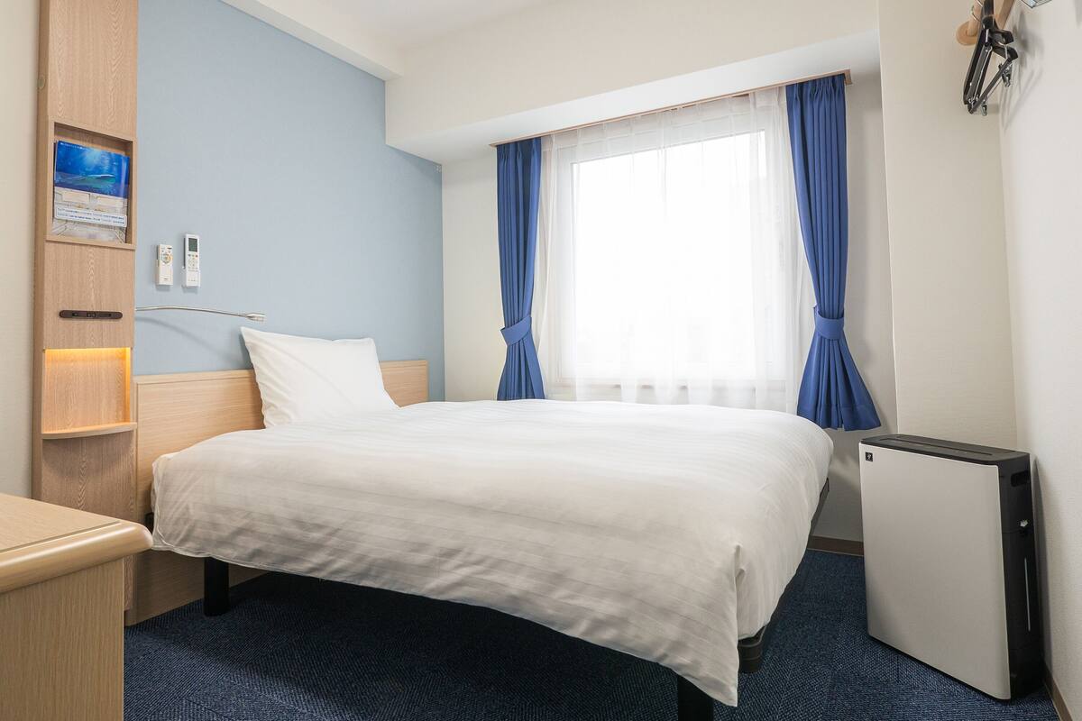 A hotel room with a bed, a desk, a small refrigerator, a nightstand, a wall-mounted lamp, a window with blue curtains, and a wall-mounted TV.