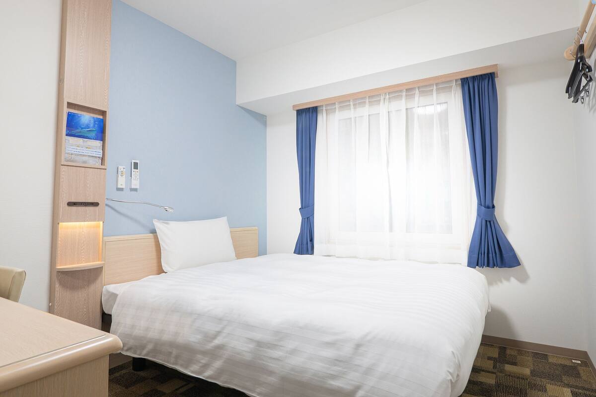 A hotel room with a bed, a desk, a chair, a nightstand, a wall-mounted lamp, and a window with blue curtains.