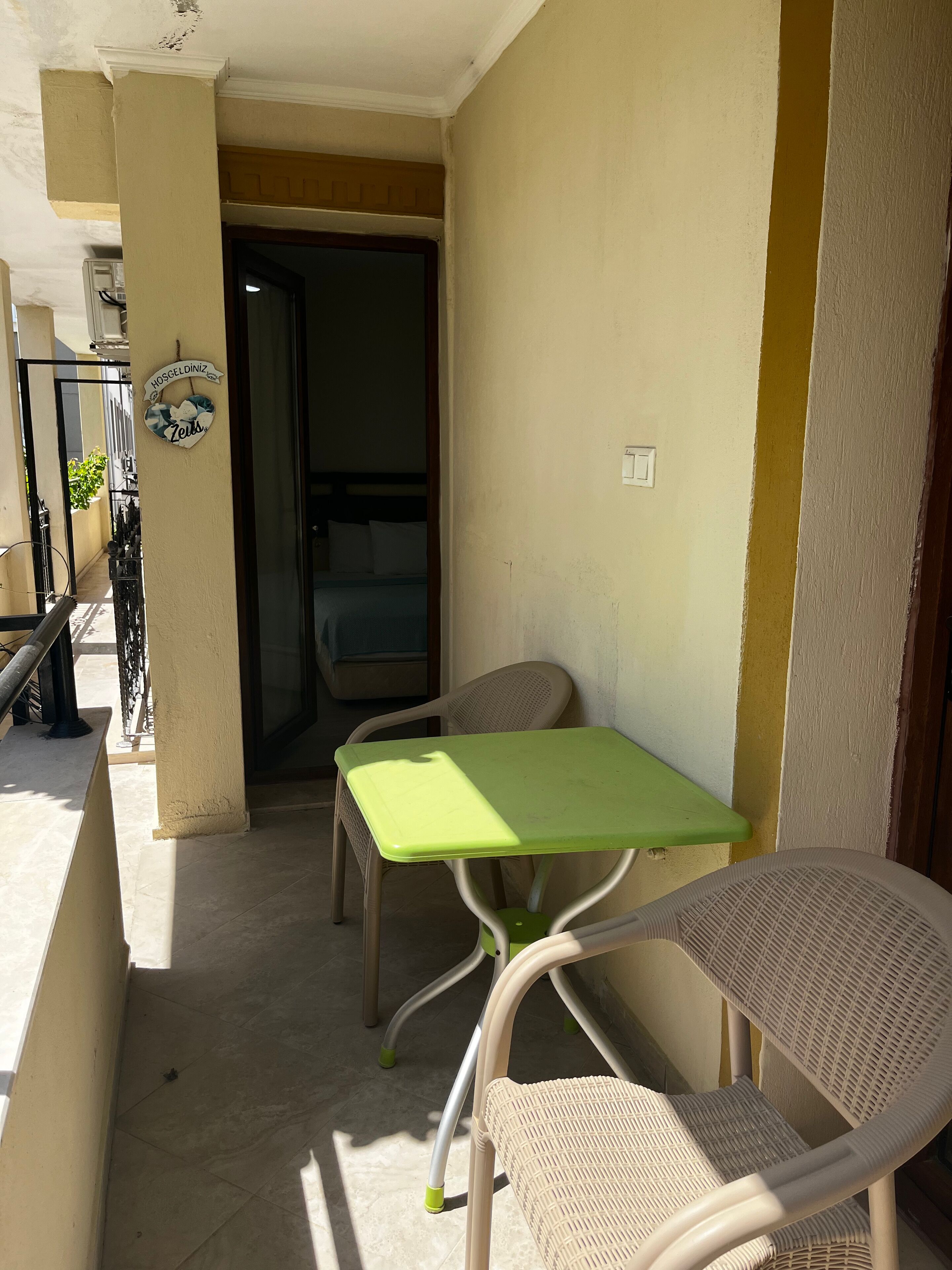 Un pequeño espacio exterior con mesa verde y sillas de mimbre, una puerta que da a una habitación y un letrero en la pared.
