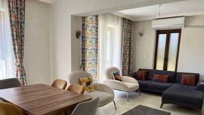 A modern living room with a wooden dining table, grey upholstered chairs, a grey sofa with orange cushions, and a black rug.