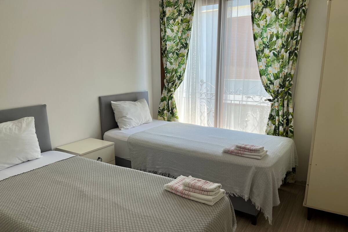 Ein Hotelzimmer mit zwei Betten, einem Nachttisch, einem Kleiderschrank und einem Fenster mit Blumenvorhängen.
