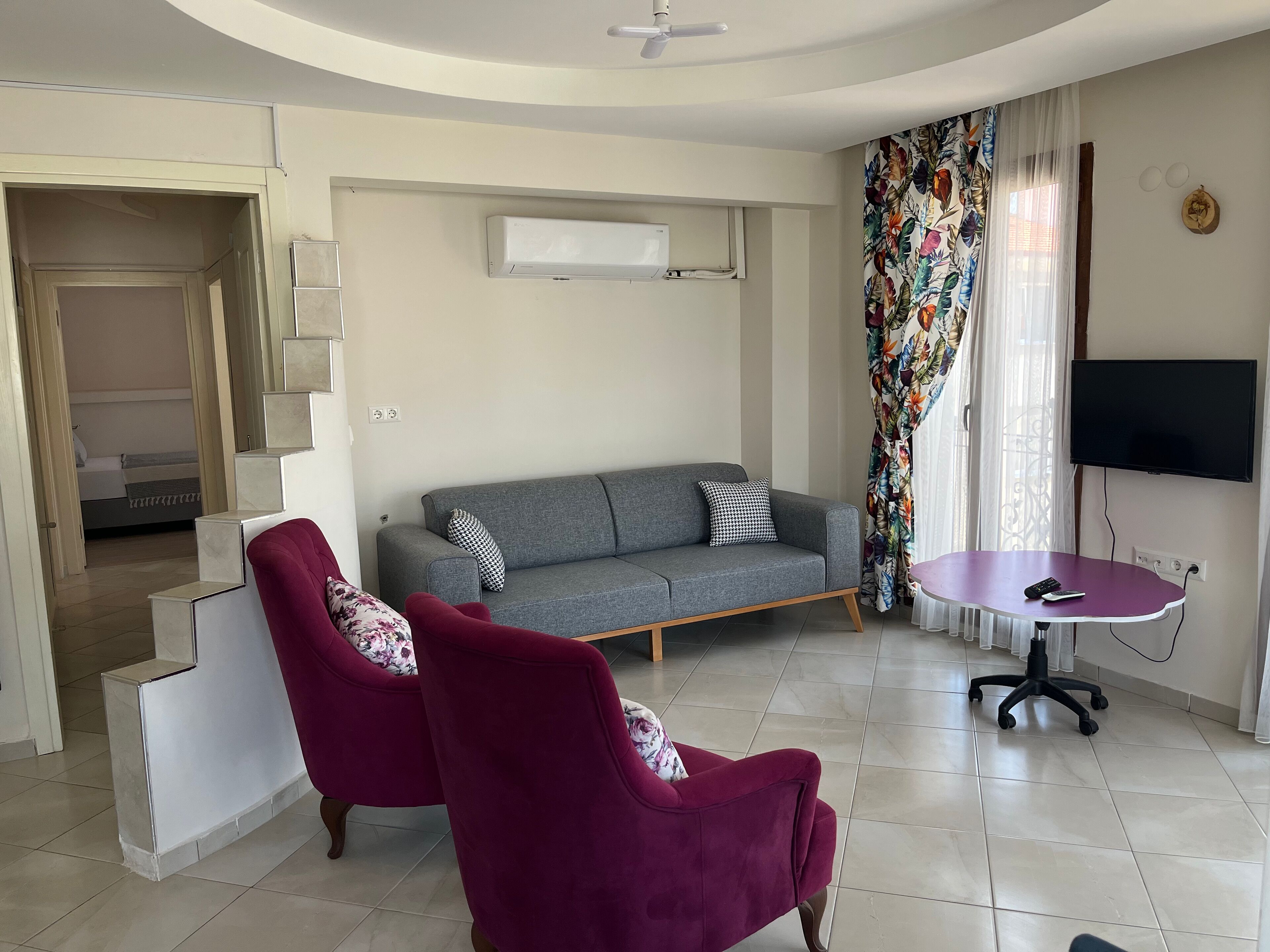 A living room with a grey sofa, two red armchairs, a small round table, and a TV on the wall.