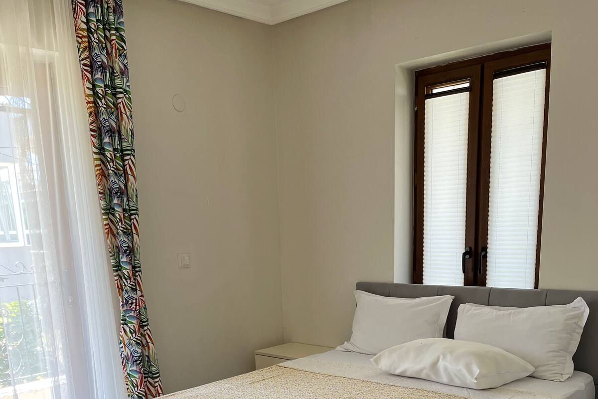 Ein Schlafzimmer mit einem Bett, einem Nachttisch, einem Fenster mit Jalousien und einem Vorhang.