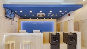 Zona de recepción de hotel con una pared azul de acento, un televisor de pantalla plana montado en la pared y dos quioscos de autoservicio.