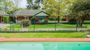 A residential property with a swimming pool, a fenced yard, and a brick house with a green roof.