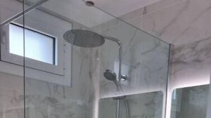 Une douche fermée par des parois de verre, avec un mur en marbre, une petite fenêtre et un meuble-lavabo noir doté de tiroirs.
