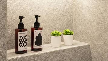 Un baño con pared de piedra, una repisa con dos botellas de champú y gel de ducha, y dos plantas en macetas.