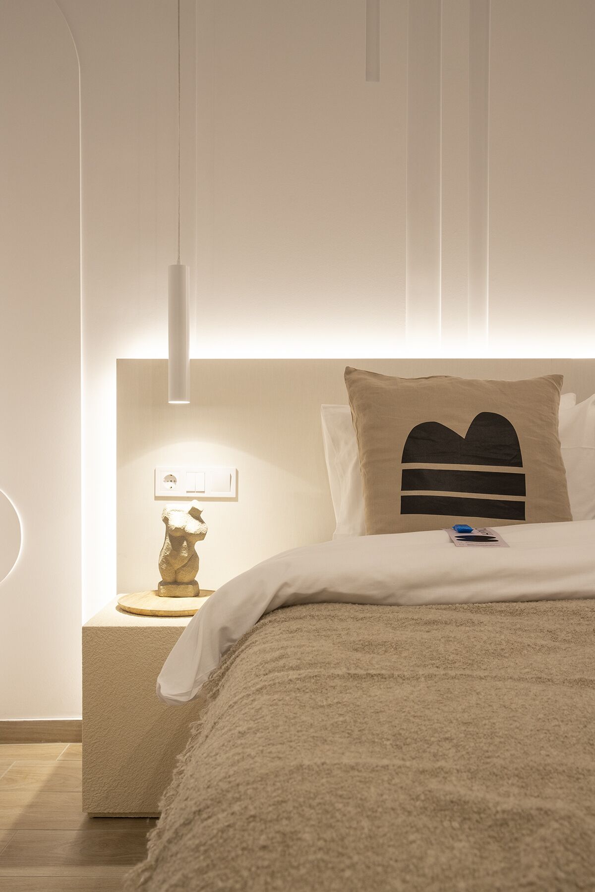 Une chambre moderne au design minimaliste, comprenant un lit recouvert d’une couverture texturée, une table de chevet avec une lampe et un luminaire fixé au mur.