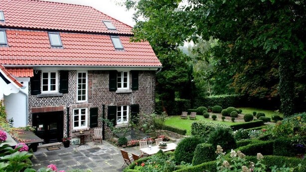 A two-story house with a red-tiled roof, brick exterior, and a well-maintained garden with hedges and flower beds. - Landhaus in Idyllischer, Ruhiger Lage und Flughafennähe (Ratingen)