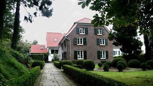 A traditional brick building with a red-tiled roof, a stone pathway leading to the entrance, and well-manicured hedges. - Landhaus in Idyllischer, Ruhiger Lage und Flughafennähe (Ratingen)