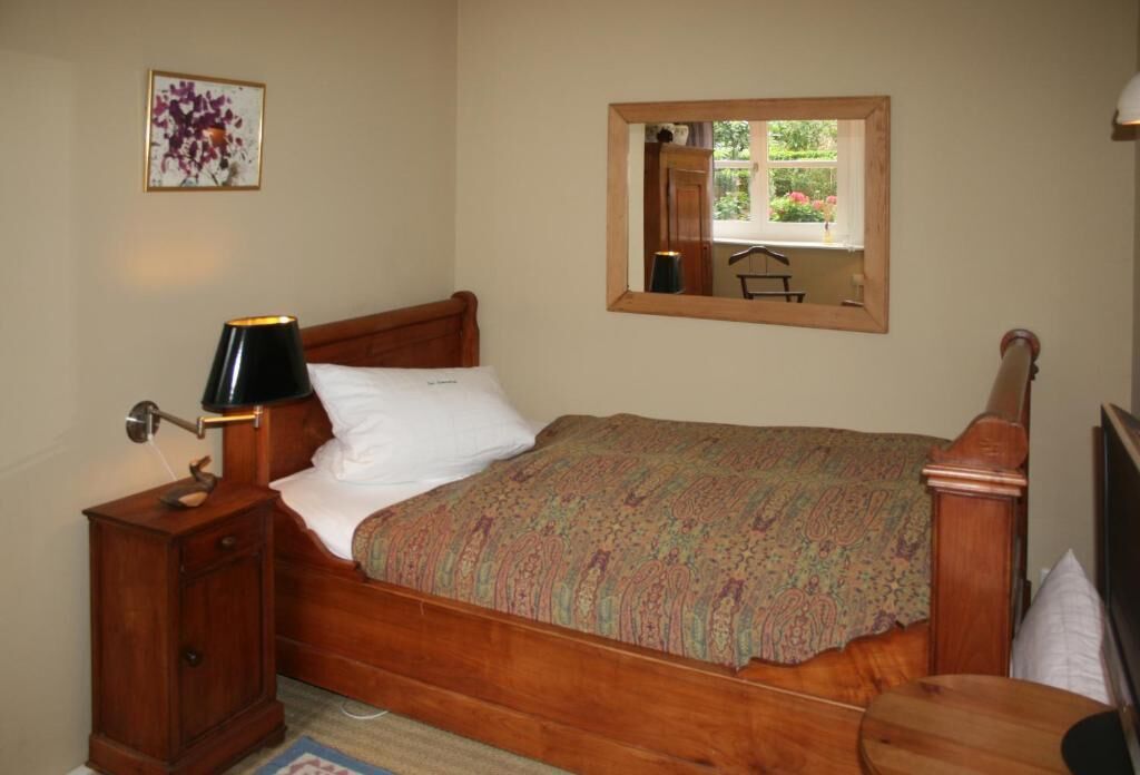 Un lit bien fait, recouvert d’une couvre-lit à motifs, une lampe de chevet, une table de nuit en bois et un miroir accroché au mur.
