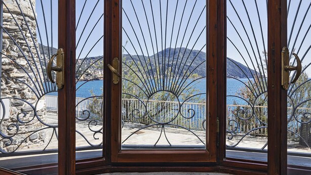 Een hoekperspectief van een kamer met een stenen muur en een metalen hekwerk dat leidt naar een balkon met uitzicht op een kustplaatsje en bergen.