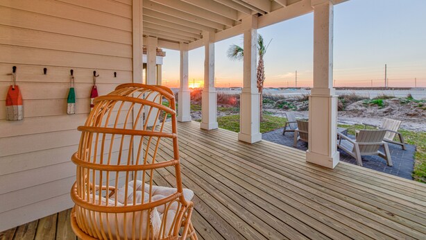Um deck de madeira com uma cadeira de vime e uma mesa, com vista para uma praia ao pôr do sol.
