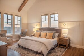 Une chambre spacieuse avec un grand lit, deux tables de chevet, un fauteuil et une vue sur la ville grâce à de grandes fenêtres.