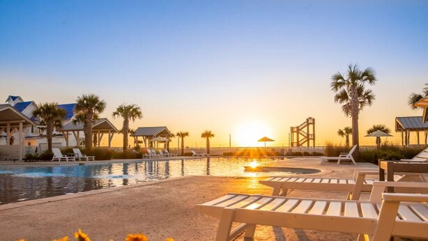 Zona de piscina con sillones de descanso y palmeras, con un atardecer al fondo y una estructura de puente.