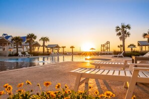 A pool area with lounge chairs and palm trees, a sunset in the background, and a bridge structure. - Gulf Views, Shared Heated Pool, Golf Cart Boardwalk to Beach (Port Aransas)