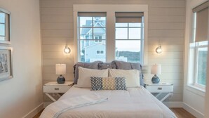 Un dormitorio con una cama grande, dos mesitas de noche con lámparas y una ventana que da a una casa.