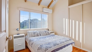 Une chambre à coucher comprenant un lit, une table de chevet, une lampe, une fenêtre avec des stores et un appareil de climatisation.