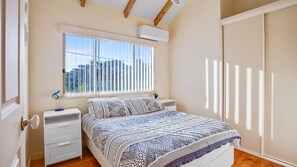 Un dormitorio con cama, mesita de noche, lámpara, ventana con persianas y aire acondicionado.