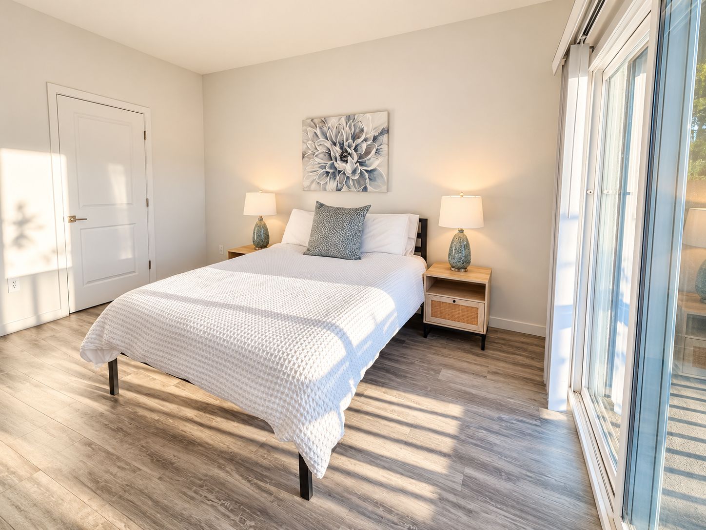 A bedroom with a bed, two bedside tables, a nightstand, a wall-mounted artwork, and a large window.