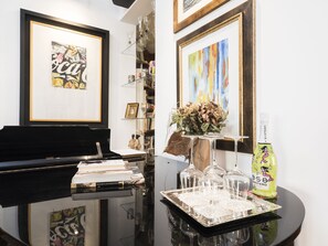A room with a piano, a glass table with books and a decorative vase, and framed artwork on the walls.