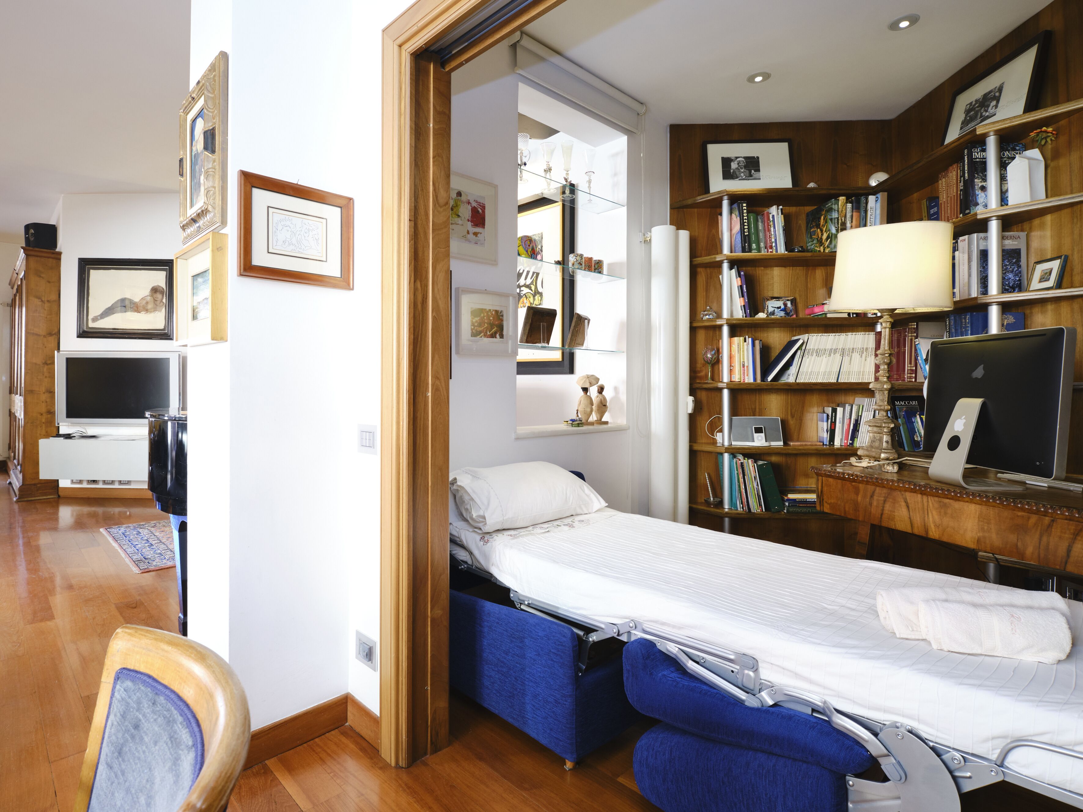 Une chambre à coucher de taille modeste, comprenant un lit, une table de chevet, une lampe et une bibliothèque remplie de livres et de photos encadrées.