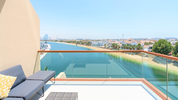 A balcony with a glass railing overlooking a body of water, a lounge area with a sofa and a small table, and a clear view of a cityscape in the distance. - Luxurious Studio With Breath-taking Palm Views (Dubai)