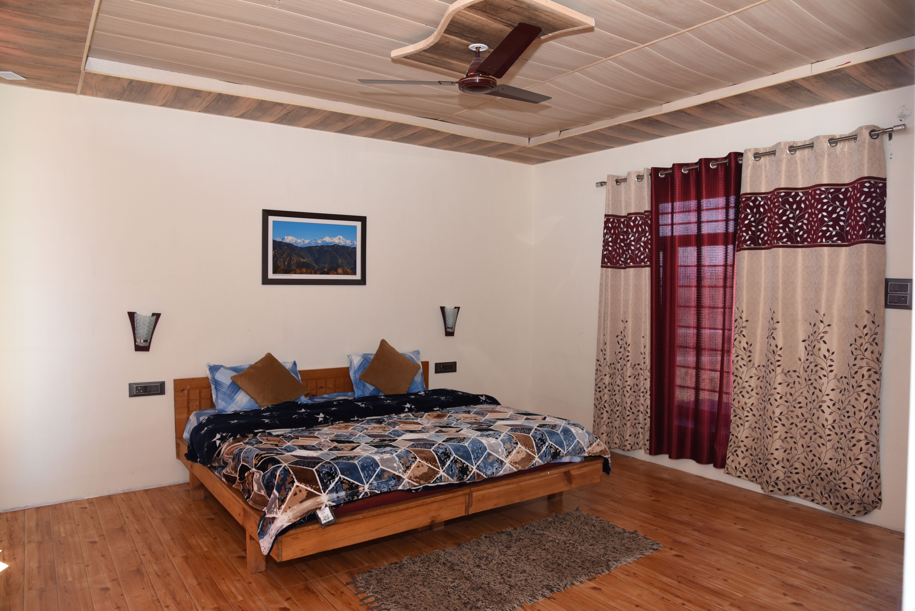 A bedroom with a wooden bed, a ceiling fan, a framed picture on the wall, and patterned curtains.