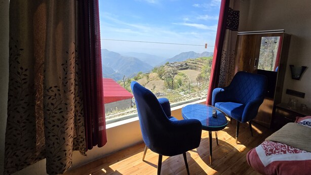 Habitación con un ventanal grande que ofrece vistas a un paisaje montañoso. Se ve una cama, una silla azul y una mesita con un control remoto.