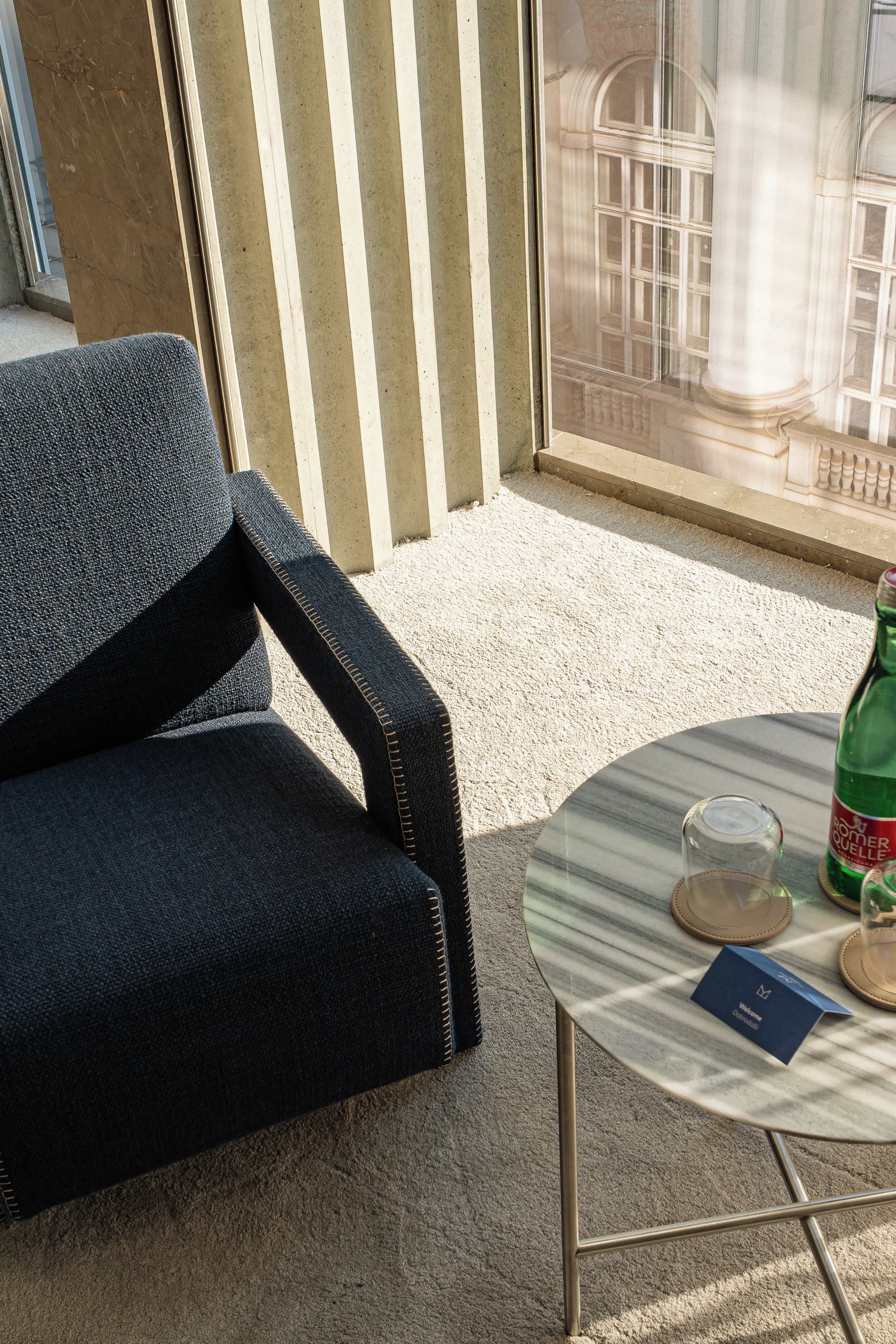 A room with a dark blue sofa, a small round table with a glass and a bottle, and a window with a view of a building.