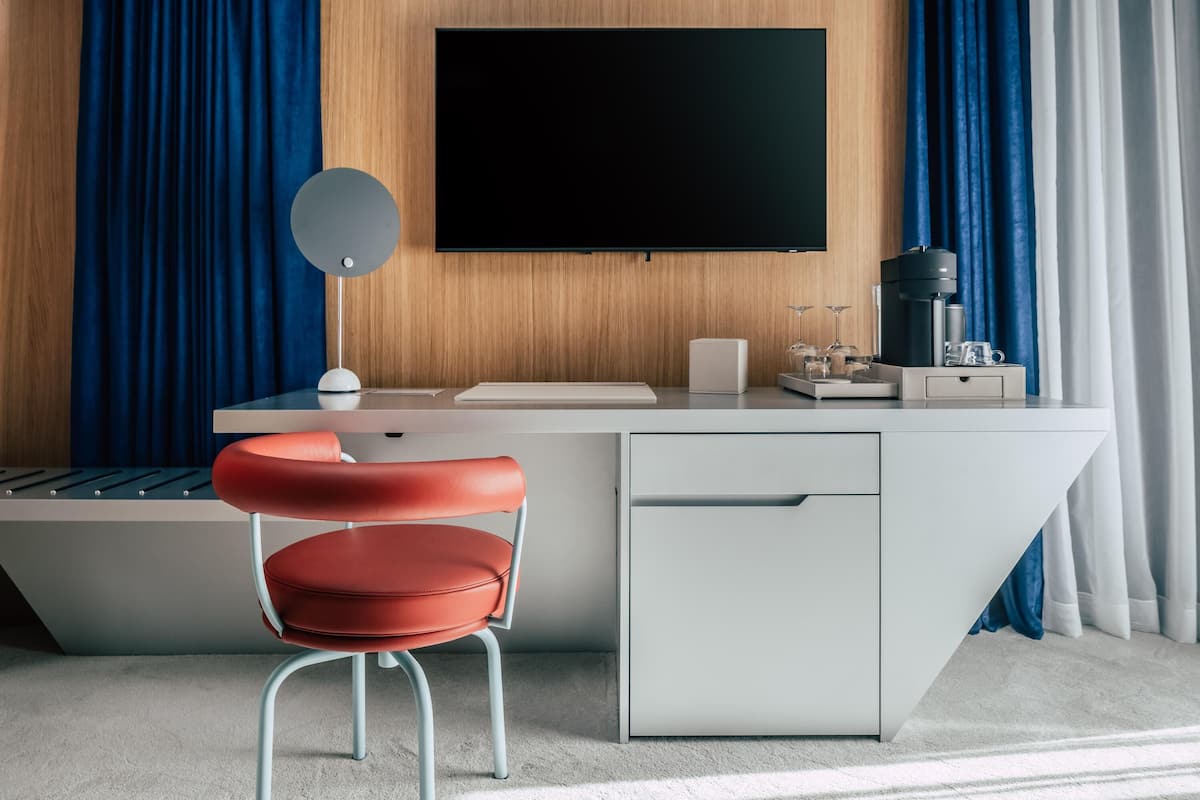 A modern hotel room with a desk, a red chair, a flat-screen TV, a coffee machine, and a minibar.