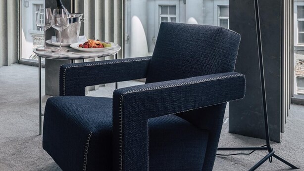 A modern lounge with a dark blue upholstered chair, a small table with a wine bottle, glasses, and fruit. A floor lamp is beside the chair. The background features a building with classical architecture and red-tiled roofs. - Met Boutique Hotel (Zagreb)