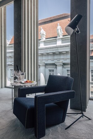 En moderne lounge med en mørkeblå polstret lænestol, et lille bord med vinflaske, glas og frugt. Der er en gulvlampe ved siden af stolen. I baggrunden ses en bygning med klassisk arkitektur og røde tegltage.