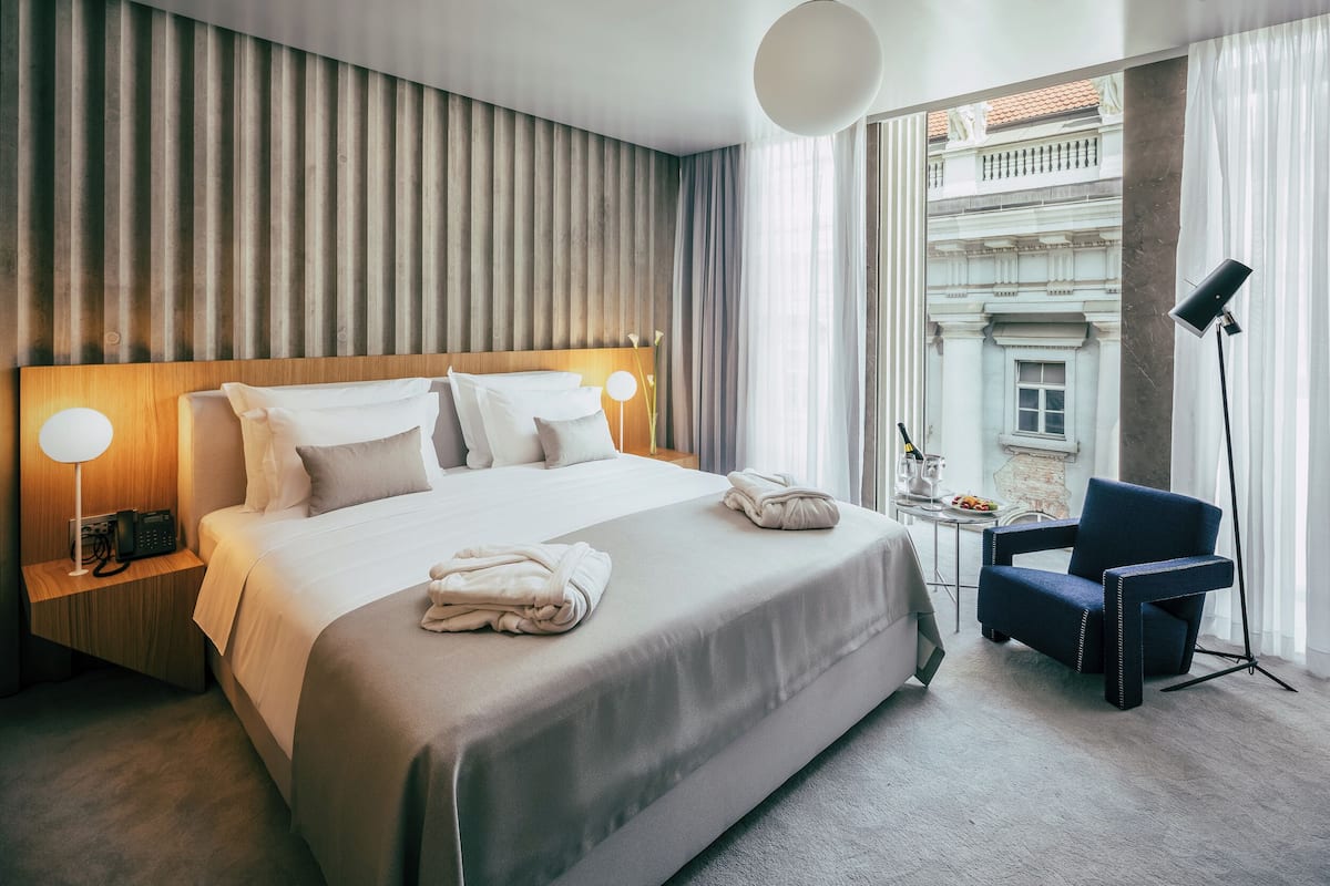 A hotel room with a large bed, bedside lamps, a chair, and a view of a building through the window.