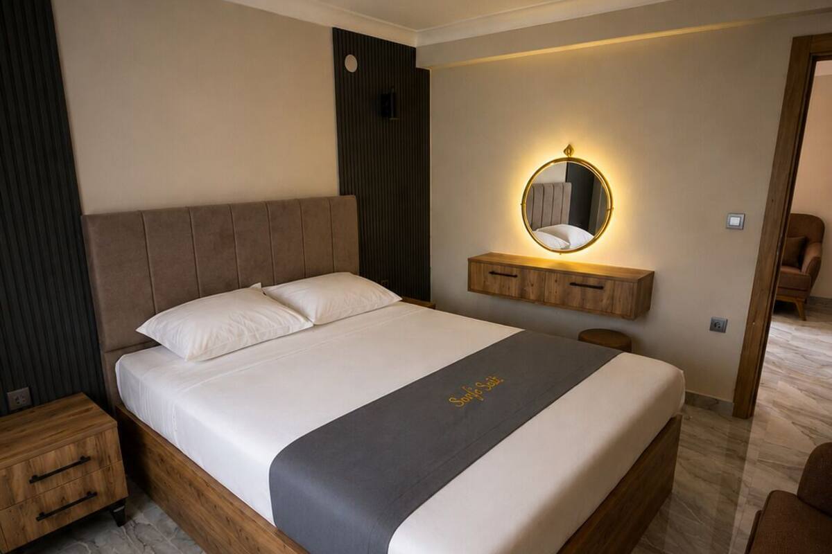 A modern bedroom with a wooden bed, a bedside table, a mirror with a light, and a dark wall panel.